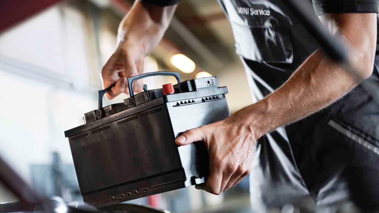 Mechanic installing a car battery under the hood in an auto repair shop. Close-up of a technician’s hands holding a black automotive battery with visible terminals, wearing a MINI Service uniform.